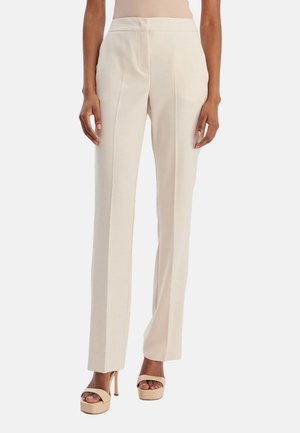 Donna che indossa pantaloni beige a vita alta e gamba dritta e sandali beige con tacco alto, in piedi su sfondo bianco.