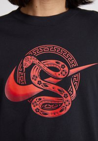 Juodas marškinėliai, kuriuose pavaizduota gyvybinga raudona grafika, vaizduojanti gyvatę, apsivyniojusią aplink stilizuotą Nike logotipą, supančius dekoratyvinio apvalaus rašto.