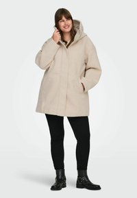 Bees värvi villasegu mantel kapuutsiga, suured ees taskud ja lõdva lõikega, kombineeritud mustade kitsaste pükste ja säärebotastega.