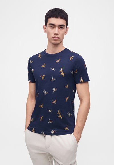 Νέος άνδρας φοράει ένα μπλε ναυτικό t-shirt με πολύχρωμα σχέδια πετώντων πουλιών και ανοιχτό μπεζ παντελόνια, στέκεται με τα χέρια στις τσέπες σε λευκό φόντο.