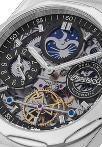 Zilveren automatische horloge met een skeletdial. Zwarte uurmarkeringen, verfijnde gouden en zilveren tandwielen, dubbele tijdsaanduiding en merklogo.
