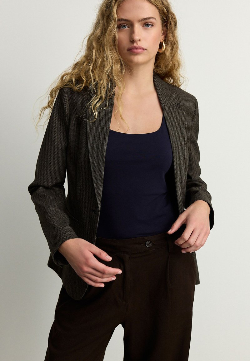 Jeune femme aux cheveux blonds bouclés porte des boucles d'oreilles créoles en or, un débardeur noir, un blazer marron foncé à carreaux et un pantalon marron foncé sur fond uni.