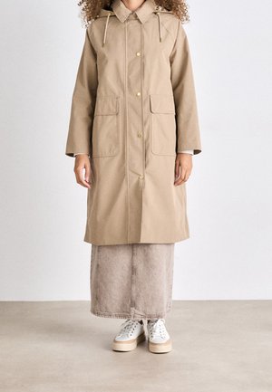 Femme portant un long manteau beige avec de grandes poches avant, une longue jupe de couleur claire et des baskets blanches à plateforme, debout sur un sol uni.