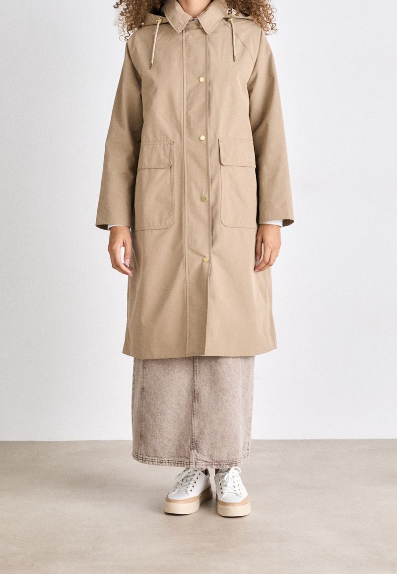 Femme portant un long manteau beige avec de grandes poches avant, une longue jupe de couleur claire et des baskets blanches à plateforme, debout sur un sol uni.