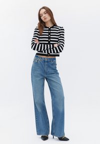 Svart- och vitrandig kort cardigan med guldknappar, kombinerad med hög midja, vida blå denimjeans och svarta spetsiga klackar.