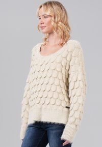 Cream-colored sweater featuring a textured design with overlapping scales, wide neckline, and ribbed cuffs and hem. Soft, fuzzy material.