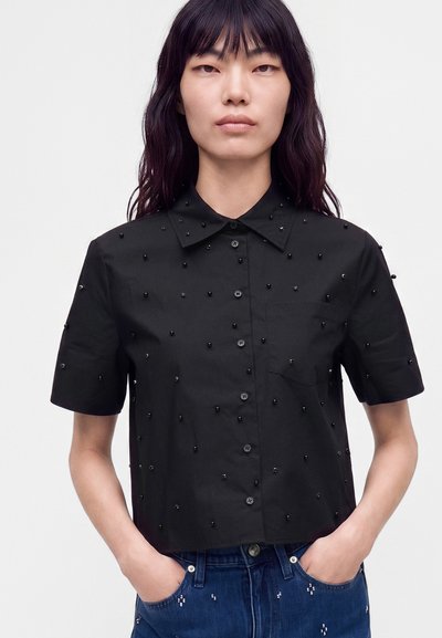 Femme aux longs cheveux foncés porte une chemise noire à manches courtes boutonnée avec de petites décorations en perles et un jean bleu avec des détails en perles.