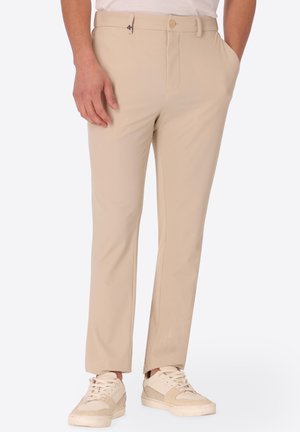 Uomo che indossa pantaloni beige a righe sottili con tasche laterali e sneaker beige con lacci, in piedi con una mano in tasca.