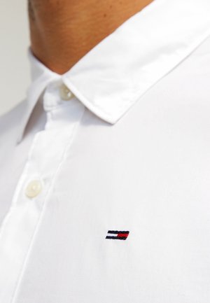 Zbliżenie osoby ubranej w białą koszulę na guziki z małym prostokątnym logo w kolorach granatowym, białym i czerwonym na piersi.