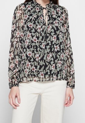Femme portant un chemisier noir transparent avec un motif de feuilles vertes et de fleurs blanches, manches longues, col à nouer, associé à un pantalon blanc.