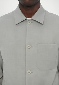 Gai pelēks pogājams krekls ar izkliedētu apkakli, priekšējo kabatu un divu pogu aizdari ar dubultšūtiem pogām. Gluda tekstūra.