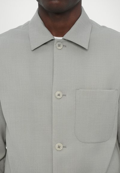 Ανοιχτό γκρί πουκάμισο με γιακά, μπροστινή τσέπη και κλείσιμο με δύο κουμπιά, με διπλοραμμένα κουμπιά. Λείος υφασμάτινος τύπος.