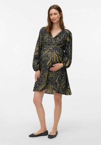 Femme enceinte debout, portant une robe à manches longues à motifs et des ballerines noires, les mains reposant doucement sur son ventre devant un fond uni.