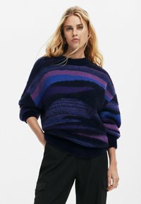 Oversized gebreide trui in tinten blauw en paars met een golvend patroon. Zachte textuur en geribbelde halslijn en manchetten. Zwarte broek gedragen.