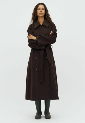 Woman with long curly hair stands with crossed arms wearing a long dark brown belted coat and black boots on a plain background.