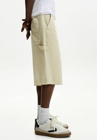 Shorts din denim bej deschis, cu un buzunar pe lateral, asortați cu adidași albi și șosete albe până la glezne. Shorts-ii au o croială lejeră și lungime până la genunchi.