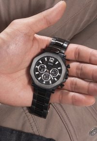 Czarny zegarek ze stali nierdzewnej z ośmiokątną tarczą, białymi znacznikami godzinowymi i sub-tarczami. Noszony na ręce na tle o teksturze.