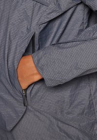 Jachetă ușoară, texturată, de culoare gri, cu un buzunar cu fermoar și detalii cu capse. Prezintă un design al mânecilor moale și ajustat.