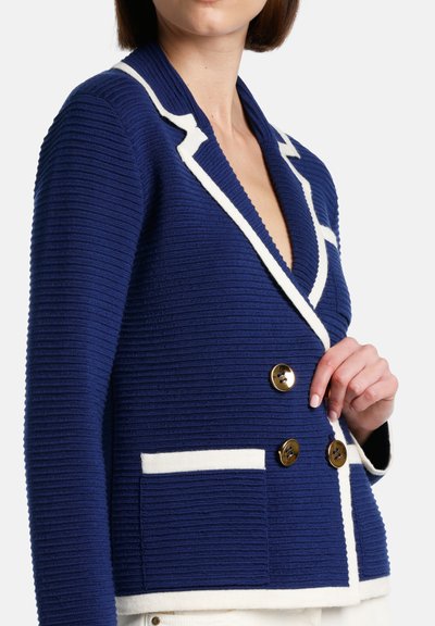 Modra teksturirana blazer jakna z kontrastnimi belimi reverji in obrobami žepov. Ima zlate gumbe in prilegajoč dizajn.