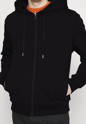 Mann trägt einen schwarzen Kapuzenpullover mit Reißverschluss und Kordeln, Hände in den vorderen Taschen, über einem orangefarbenen Rundhalsshirt.