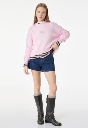 Młoda kobieta ubrana w różowy sweter z napisem „Angel”, pod spodem w paski koszulę, jeansowe szorty, szare skarpetki i czarne kozaki za kolano.