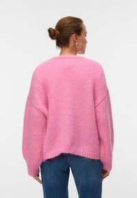 Γυναίκα με τα μαλλιά μαζεμένα σε κότσο, φοράει ένα φαρδύ, αφράτο ροζ πουλόβερ και μπλε τζιν, με την πλάτη στραμμένη σε ένα απλό λευκό φόντο.