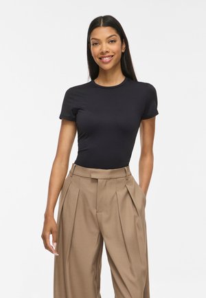 Jeune femme aux longs cheveux foncés portant un t-shirt noir ajusté à manches courtes et un pantalon large plissé beige taille haute, debout sur un fond blanc.
