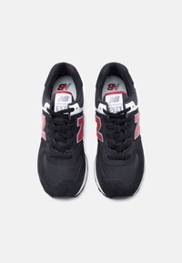 Ζευγάρι μαύρα αθλητικά παπούτσια New Balance 574 με κόκκινες λογότυπες "N", λευκές λεπτομέρειες και μαύρα κορδόνια, σε προβολή από πάνω, πάνω σε λευκό φόντο.