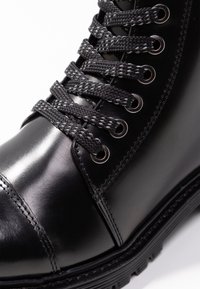 Primer plano de una bota de cuero negro con cordones gruesos de patrón negro y gris pasados por ojales de metal.