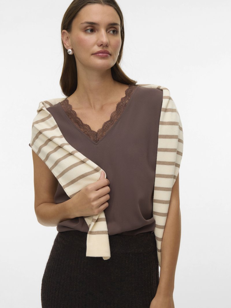 Γυναίκα που φοράει μπλουζάκι V-λαιμόκοψη με καφέ δαντέλα, σκούρα φούστα ψηλόμεση και κρεμ πουλόβερ με ανοιχτό καφέ ρίγες στους ώμους.