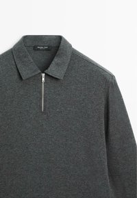 Grijze polo met rits en platte kraag, gemaakt van textuurstof. Bevat een metalen rits en subtiele stikdetails op de schouder.
