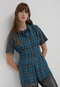 Camicia a quadri color teal con finitura testurizzata, caratterizzata da un colletto, maniche corte grigie e linee nere e bianche a contrasto.