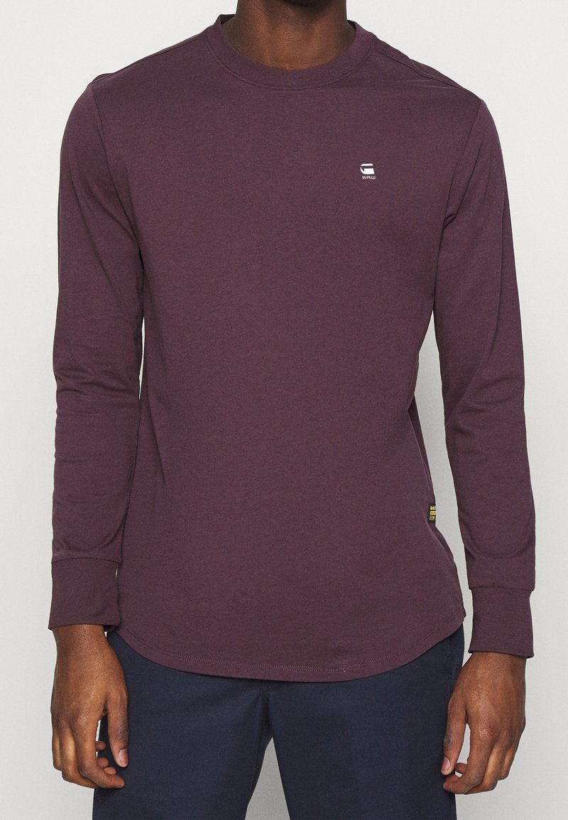 Homme portant une chemise bordeaux ajustée à manches longues avec un petit logo blanc sur la poitrine et un pantalon sombre, mains détendues sur les hanches.