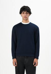 Un joven con cabello rizado y oscuro lleva un sudadera azul marino sobre una camiseta blanca y pantalones negros, de pie contra un fondo blanco liso.