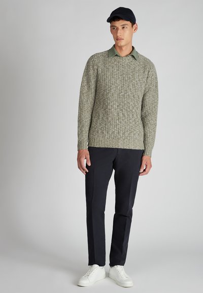 Jonge man in een groene gebreide trui over een colbert, zwarte broek, witte sneakers en een zwarte pet, staand tegen een neutrale achtergrond.