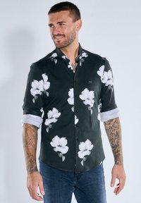 Schwarzes Hemd mit weißem Blumenmuster, mit Knopfkragen, hochgekrempelten Ärmeln, aus einem Stoff mit glatter Textur, getragen mit blauen Jeans.