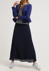 Donkerblauwe geplooide rok gecombineerd met een navy jasje en een gestreept topje; lichte textuur; geaccentueerd door grijze sneakers.