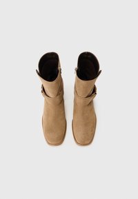 Bottes de cheville en daim beige avec un bout carré, des sangles enroulées et une fermeture éclair sur le côté. Texture lisse et détails de design minimalistes.