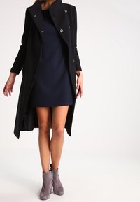 Manteau long noir avec de grands revers et des accents de boutons, porté sur une robe bleue marine. Bottines grises à la cheville avec une texture douce complètent le look.