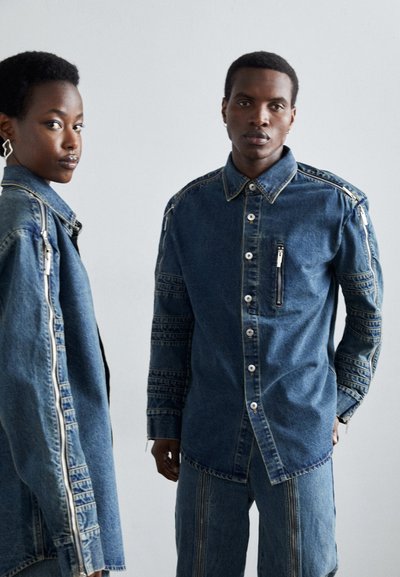 Cămașă din denim cu închidere cu butoane, având un buzunar cu fermoar și accente cu model pe mâneci. Culoare albastru deschis, croială lejeră.