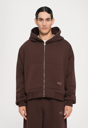 Brown zip-up hoodie in soft fabric with large pockets, oversized fit, and a hood. Features subtle logo accent on the front.