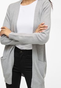 Hellgraue Strickjacke aus weichem Stoff, offene Front, mit gerippten Bündchen und zwei vorderen Taschen, getragen über einem weißen Oberteil und schwarzen Hosen.
