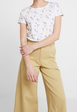 Kobieta ubrana w biały top z krótkim rękawem z drobnym kwiatowym nadrukiem i szerokie beżowe spodnie z wysokim stanem, z założonymi na piersiach rękami.