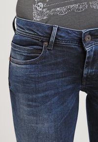 Dark blue denim jeans with a slim fit, featuring a classic five-pocket design, metal rivets, and subtle fading along the thighs.