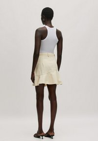 Witte tanktop en een crèmekleurige, uitlopende minirok. De rok heeft een gebogen zoom en subtiele textuur. Draagt een paar zwarte open-teen hakken.