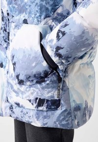 Veste gonflante avec un motif abstrait de montagnes bleues et blanches, poches latérales zippées et une étiquette de marque avec "GUESS EST. 1981."