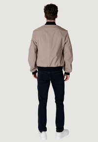Giacca bomber beige con colletto e polsini neri, caratterizzata da una texture liscia e vita elasticizzata, abbinata a jeans in denim scuro e scarpe da ginnastica bianche.