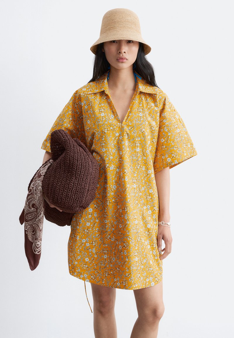 Femme portant une robe jaune à fleurs à manches courtes et un chapeau seau beige, tenant un sac en tricot marron avec une écharpe à motifs.