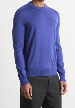 Man draagt een nauwsluitende kobaltblauwe trui met lange mouwen, geribbelde manchetten en zoom, gecombineerd met een zwarte broek.