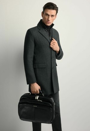 Czarna skórzana teczka z wieloma zamykanymi na suwak przegródkami, trzymana przez mężczyznę w ciemnoszarym wełnianym płaszczu o wyraźnej konstrukcji, stojącego na jasnym tle.